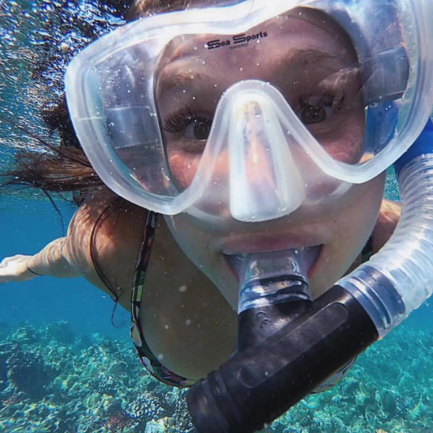 A close-up of a snorkeler underwater, wearing a snorkel mask and smiling while swimming in clear blue water with corals visible below.