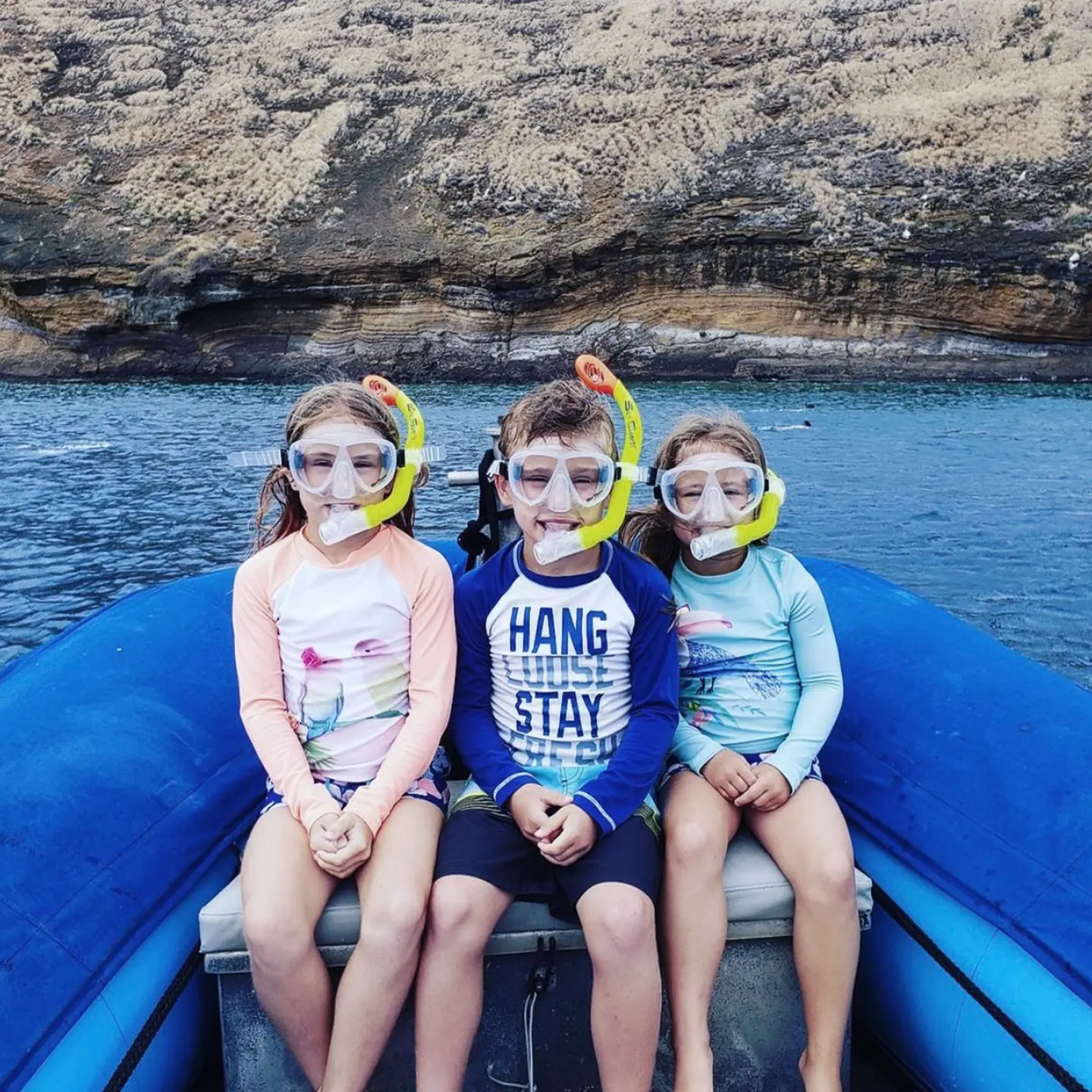 Three children wearing snorkel masks sitting together on a boat, smiling with a scenic rocky coastline in the background.