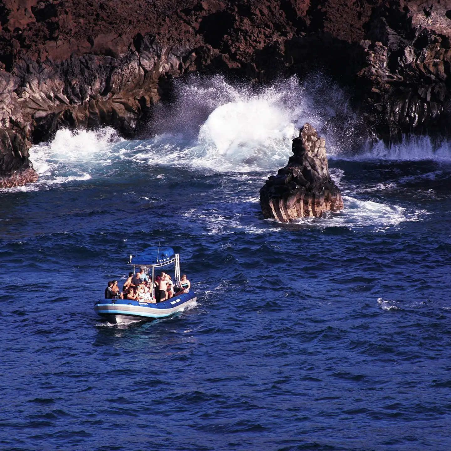 A boat filled with passengers navigating near dramatic rocky cliffs with waves crashing into the rocks, surrounded by deep blue ocean water.