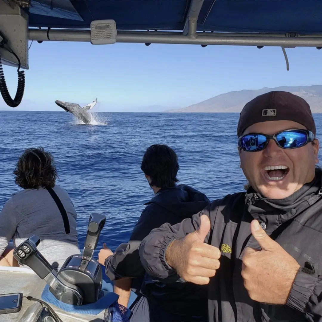 Excited boat passengers watch a humpback whale breaching against a scenic ocean backdrop